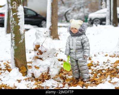 Ein lustiges Mädchen in einem silbernen Jumpsuit hält eine Spielzeugschaufel und modelliert einen Schneemann. Stockfoto