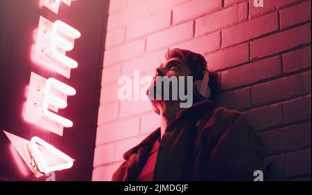 Setzen Sie Ihre Kopfhörer auf und vergessen Sie alles andere. Ein junger Mann, der Kopfhörer trägt, während er sich nachts an ein Gebäude lehnt. Stockfoto