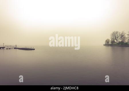 Ein Bootsdock und eine einsame Insel im Nebel, die bei Sonnenaufgang tief auf dem Wasser eines großen Flusses sank Stockfoto