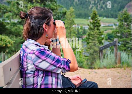 Reife Frau, die auf der Bank sitzt und ein Fernglas benutzt Stockfoto