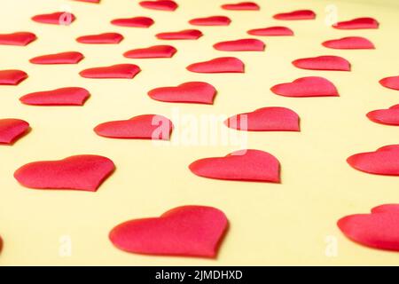 Viele identische rote Seidenherzen liegen versetzt auf einem gelben Hintergrund. Symbol der Liebe, Zärtlichkeit und Leidenschaft Stockfoto