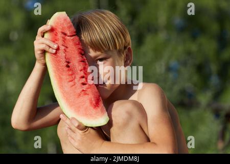 Kleiner Junge, der ein Stück Wassermelone vor dem Gesicht auf grünem Naturhintergrund hält. Stockfoto