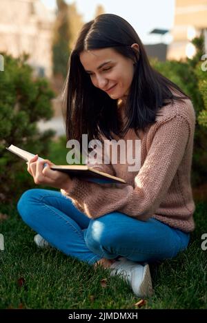 Junge Frau, die auf Gras sitzt und ein Buch auf den Knien hat Stockfoto
