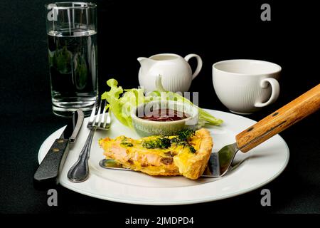 Eine Portion frisch gebackener Brokkoli-Lachs-Quiche in eine weiße Keramikschale auf schwarzem Hintergrund geben. Stockfoto