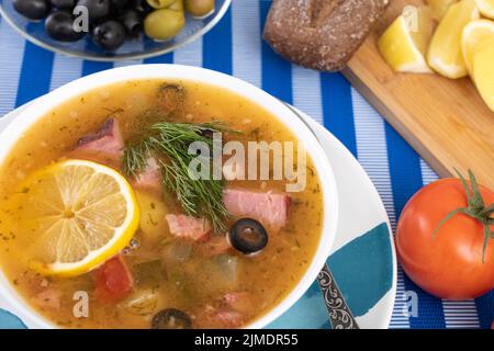 Suppe mit Fleisch, Wurst, gesalzene Gurken auf wite Schüssel. Schwarzbrot, Zitronen, Oliven, Tomaten auf Farbe blau Hintergrund. Stockfoto