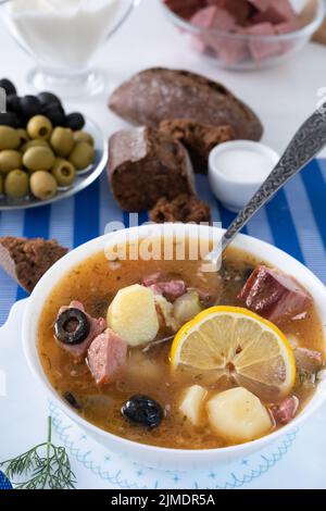 Traditionelle russische Suppe Soljanka mit Fleisch, Würstchen, gesalzene Gurken auf einer Schüssel gekocht. Schwarzes Brot, Zitrone, Olive und Toma Stockfoto