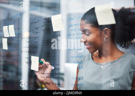 Glückliche, inspirierte und selbstbewusste Geschäftsfrau, die Ideen zum Brainstorming auf einer transparenten Glasscheibe mit Haftnotizen schrieb. Starker Unternehmer und CEO Stockfoto