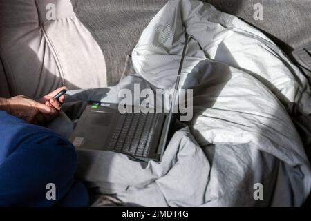 Ein Mann, der auf einem unordentlichen, ungefertigten Bett sitzt und an einem Laptop arbeitet Stockfoto