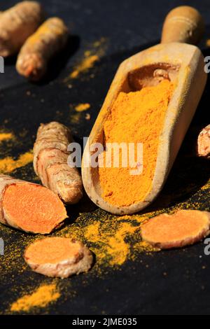 Kurkuma Pulver in einem Holzlöffel und Wurzeln auf einem dunklen Tisch (Curcuma longa), Kurkuma Stockfoto