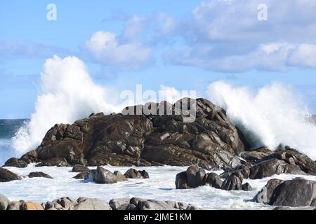 Wolken berühren das Meer und verschmelzen Weiß, Blau und Grau. Felsen und Gezeitenbecken im Vordergrund spiegeln den großen Felsen und den Schaum des Brennpunktes wider. Stockfoto