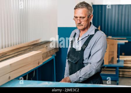 Porträt eines älteren Zimmermanns oder Zimmermanns, der mit Holzbrettern in einer Tischlerwerkstatt arbeitet. Stockfoto