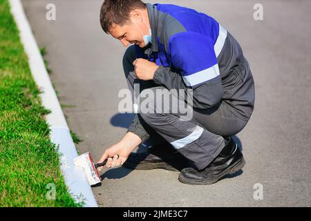 Ein Arbeiter in Overalls malt an einem Sommertag die Bordsteinkante mit weißer Farbe. Städtischer Dienst, Verbesserung der Territorien. Stockfoto