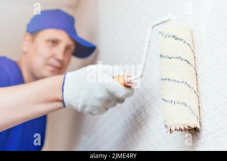 Ein Maler mit Mütze und Handschuhen bemalt die Wand mit einer Farbrolle. Porträt eines jungen Arbeiters in Uniform. Stockfoto