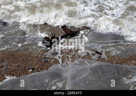 Die verstümmelte Hinterachse eines Autos, das am Strand gestrandet ist und von der Flut überspült wird. Stockfoto