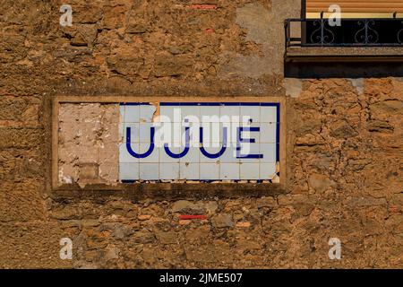 Altes, beschädigtes Fliesenschild mit dem Dorfnamen auf einer Steinmauer in einem malerischen mittelalterlichen Dorf Ujue im Baskenland, Navarra, Spanien Stockfoto