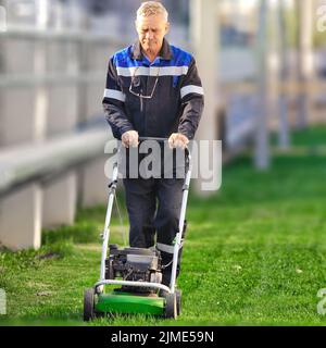 Ein älterer Arbeiter mäht an einem sonnigen Tag den Rasen. Ein Mann mit Arbeitskleidung arbeitet mit einem Rasenmäher auf einer grünen Wiese. Stockfoto