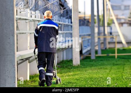 Ein älterer Mann in Arbeitskleidung geht an einem sonnigen Tag mit einem Rasenmäher. Ein Rentner arbeitet in Teilzeit. Rückansicht Stockfoto