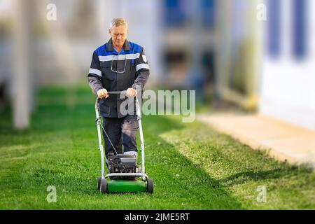 Ein älterer Arbeiter mäht an einem sonnigen Tag den Rasen. Ein Mann mit Arbeitskleidung arbeitet mit einem Rasenmäher auf einer grünen Wiese. Stockfoto