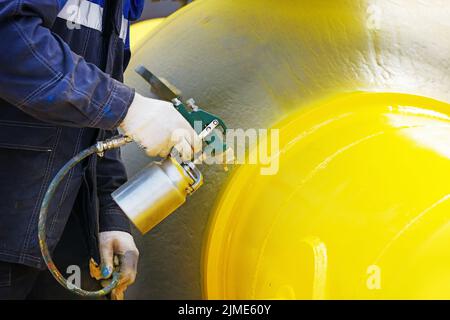 Ein Maler in Arbeitskleidung malt an einem Sommertag ein Absperrventil aus Metall zur Vergasung aus einer Kompressorpistole. Stockfoto