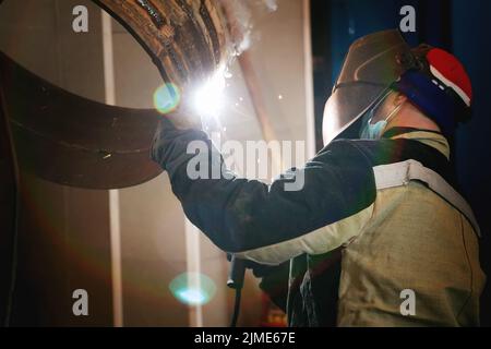 Dies ist ein Schweißer in einer Schutzmaske und Arbeitskleidung bei der Arbeit in der Produktionshalle. Eine echte Arbeitsumgebung. Stockfoto