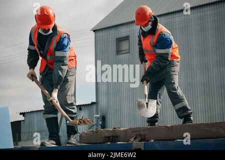 Zwei Arbeiter in Schutzmützen, Arbeitskleidung und einer medizinischen Maske arbeiten auf einer Baustelle mit Schaufeln. Harte körperliche Arbeit. Stockfoto