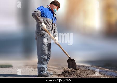 Ein Arbeiter in einer medizinischen Maske und Arbeitskleidung steht mit einer Schaufel auf einem verschwommenen Hintergrund. Körperliche Arbeit. Stockfoto