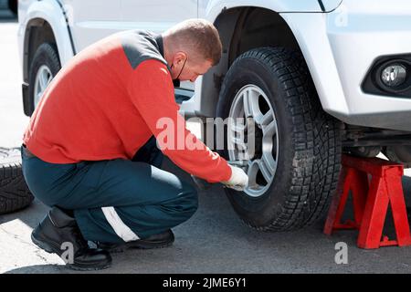 Ein Automechaniker in einer medizinischen Maske wechselt an einem sonnigen Tag auf der Straße ein Rad an einem Auto. Hilfe unterwegs Stockfoto