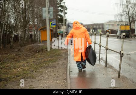 Arbeiter entfernt Müll von der Straße. Mann im orangen Regen. Schwarze Tasche in der Hand. Straßenarbeiter. Stockfoto