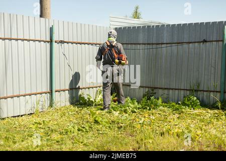 Mann mäht Gras in der Nähe des Zauns. Mann schneidet Rasen. Guy arbeitet als Rasenmäher. Stockfoto