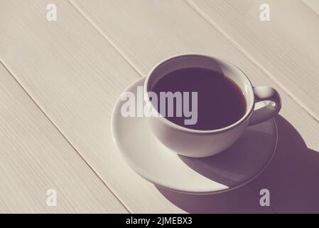 Schwarzer Kaffee in weißer Tasse mit Untertasse auf hellem Holztisch, Schatten, Kopierraum Stockfoto