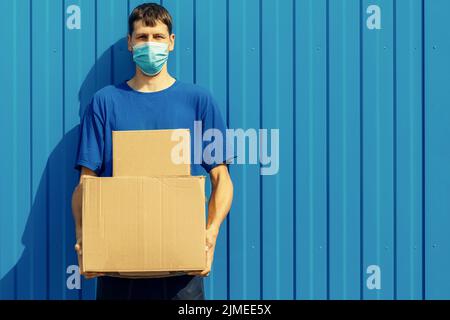 Ein junger kaukasischer Kurier, der freiwillig in einer medizinischen Maske arbeitet, hält Pappkartons in seinen Händen. Stockfoto