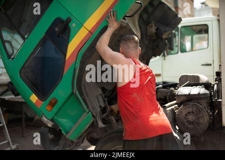 Fahrer repariert Stapler. Der Fahrer hebt die Kabine des Staplers an. Großes Auto. Typ in rotem T-Shirt. Stockfoto