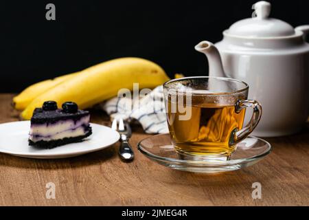 Blick auf heißen Tee in transparenter Tasse und Scheibe Blaubeer-Käsekuchen in weißer Keramikschale. Stockfoto