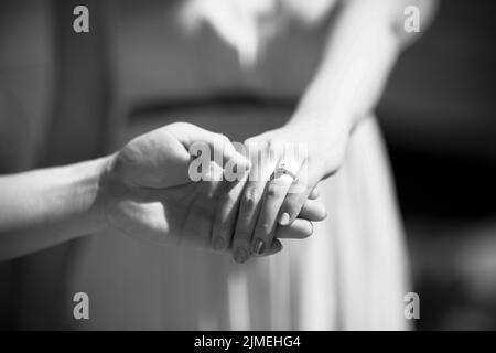 Eine Nahaufnahme eines Bräutigams, der die Hand der Braut mit einem schönen Ring an ihrem Finger in Graustufen hält Stockfoto