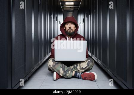 Der Hacker in einer Haube sitzt mit dem Computer unter den Servern, der Einbrecher. Speicherung von Daten, cloudy Services, Systemausbruch, attac Stockfoto