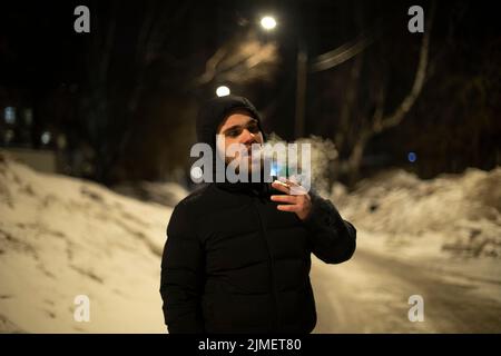 Kerl raucht Zigarette auf der Straße. Mann in schwarzer Jacke. Mann in schwarzer Kapuze. Rauchen ist gesundheitsschädlich. Unbekannte Person. Gefährliche Person. Stockfoto