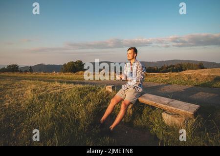 Fotograf in einem Hemd, das auf einem Hügel sitzt, bedient seine Drohne, um Fotos von der Landschaft unten zu machen. Technologische Fortschritte bei Videofilmen. Stockfoto