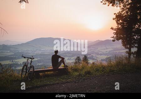 Aktiver Athlet, der auf einer Bank sitzt, nachdem er auf dem Fahrrad auf die Spitze eines Hügels gegangen ist und den Sonnenuntergang beobachtet hat. Beskiden, Tschechische republik, Herz der EU Stockfoto