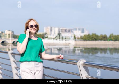 Ein lächelndes Mädchen in Sonnenbrillen, das mit Freunden telefoniert, gestikiert, am Flussufer in der Stadt ein Date macht. Mädchen läuft am Stadtufer entlang Stockfoto