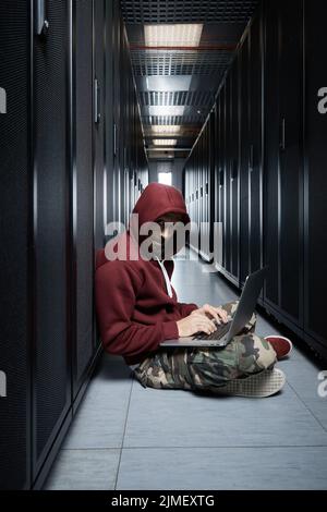 Der Hacker in einer Haube sitzt mit dem Computer unter den Servern, der Einbrecher, Gesicht mit einer Haube bedeckt. Speicherung von Daten, trüber Service Stockfoto