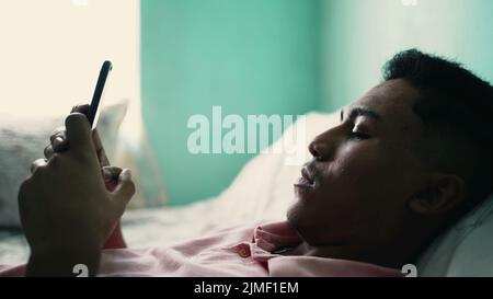 Ein hispanischer Mann, der das Mobiltelefon im Bett liegend ansieht und mit ernsthaftem Ausdruck auf den Bildschirm starrt Stockfoto