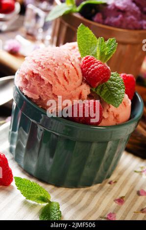 Rosafarbene Himbeer-Eiscreme-Kugeln in Tonschüsseln auf einem hölzernen Küchentisch mit Beeren und Löffeln zur Seite Stockfoto