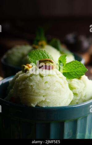 Pistazien-Eiscreme-Kugeln in Tonschüsseln auf einem hölzernen Küchentisch mit Krümel und Nüssen zur Seite Stockfoto