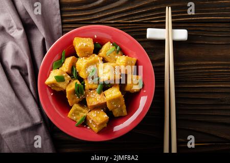 Flach legen Blick auf knusprig tief rühren gebratenen Tofu Würfel mit Schnittlauch in Tonschale auf hölzernen Küchentisch mit Serviette beiseite Stockfoto