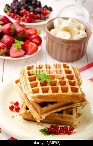 Stapel von drei belgischen Waffeln auf weißem Küchentisch mit Erdbeeren, Bananen und roten Johannisbeeren Stockfoto