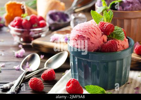 Rosafarbene Himbeer-Eiscreme-Kugeln in Tonschüsseln auf einem hölzernen Küchentisch mit Beeren und Löffeln zur Seite Stockfoto