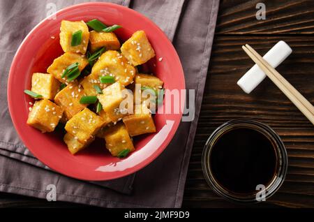 Flach Lay Blick auf knusprig frittierte Tofu-Würfel mit Schnittlauch in Tonform auf Holzküchentisch mit Serviette und Sojasauce als Stockfoto