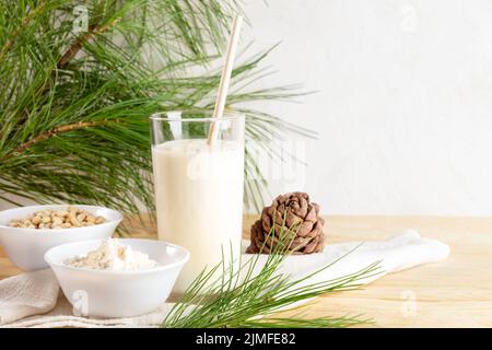 Zedernmilch im Glas und Zutaten dafür Kochmehl und Nüsse, Kegel, Zweige auf Serviette auf Holztisch. Pflanzliche, nicht milchfreie Alternativmilch. O Stockfoto