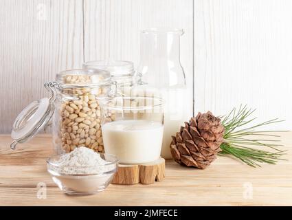 Glas und Flasche mit veganer Zedernmilch, Mehl in der Schüssel und Nüsse im Glas, Kegel, Zweig auf Holztisch. Pflanzliche Alternativmilch. Laktosefrei Stockfoto