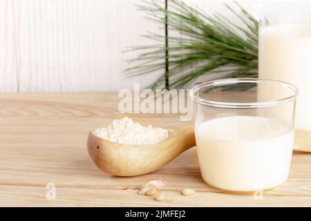 Bio-Milchaustauschstoff. Kleines Glas mit Zedernmilch und Mehl in Holzlöffel zum Kochen auf Holztisch aus nächster Nähe. Pflanzliche Alternativmilch. L Stockfoto